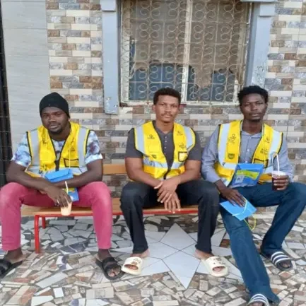 Trois jeunes bénéficiaires en gilet de travail assis devant le local du projet Avenir Volaille